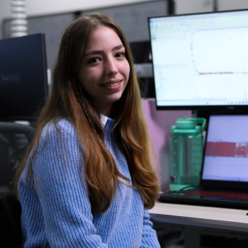 Young woman with long brown hair wearing a blue sweater, smiling at her desk in an office with computer screens displaying technical designs.